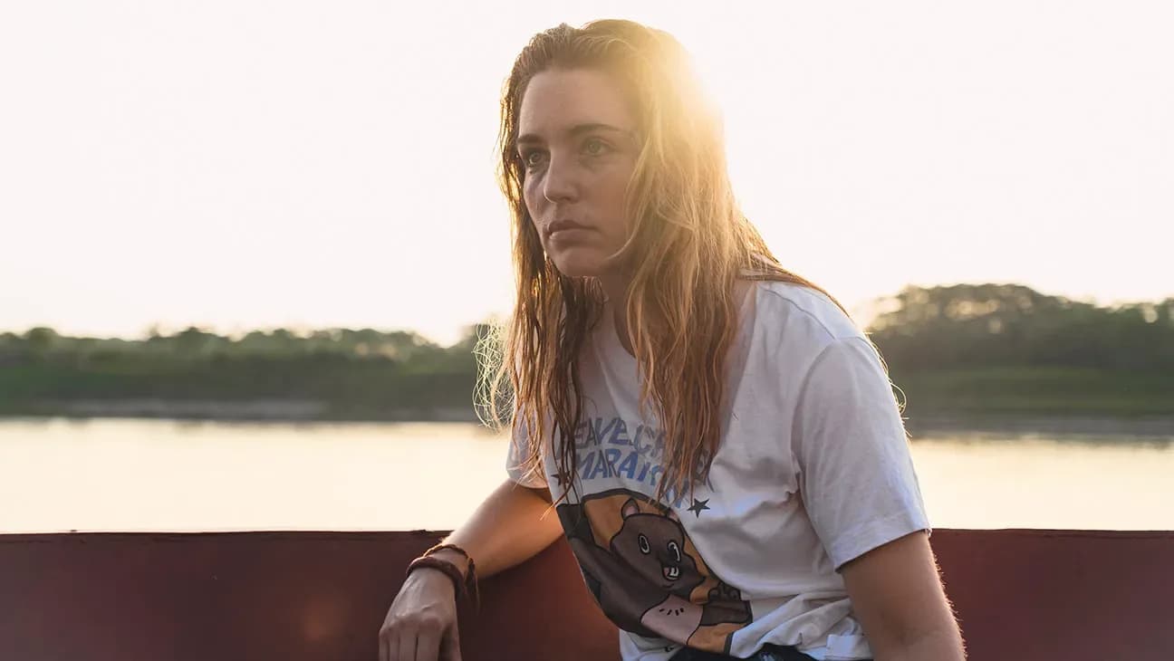 Jessica Rothe in a tense scene from 'Beware Boiúna', standing near the Amazon River with a look of fear.