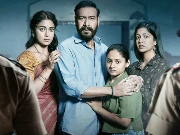 Ajay Devgn in a tense scene from Drishyam: The Conclusion, standing against a dimly lit backdrop with a determined expression.