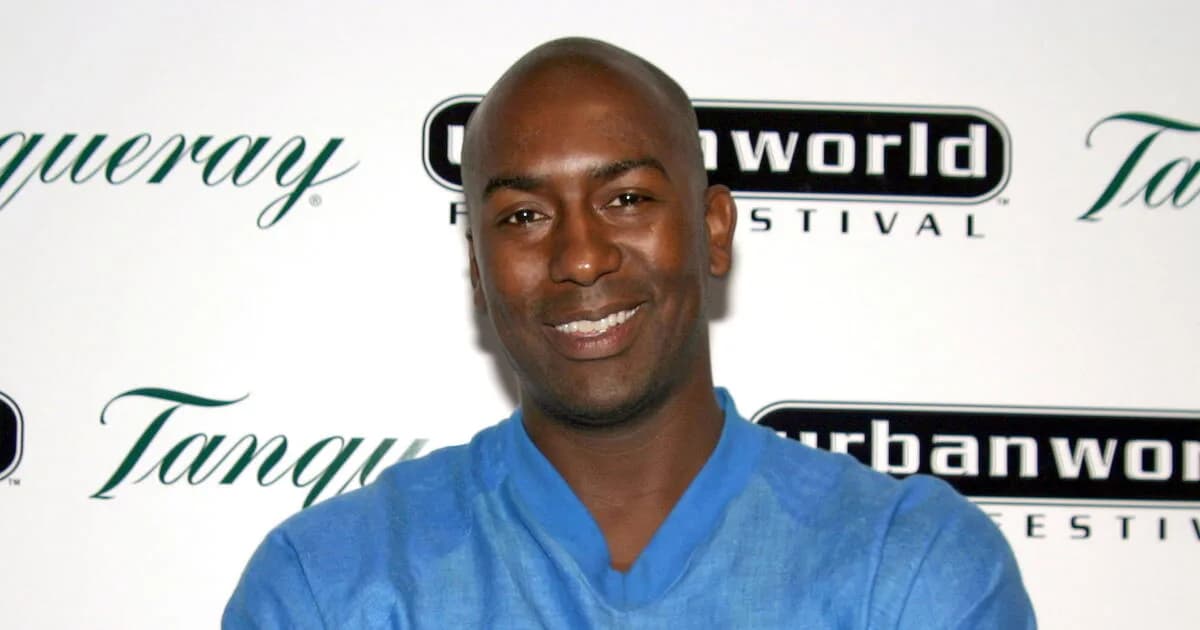 A black-and-white photo of Reggie McFadden in a suit, mid-laugh, standing in front of a microphone.