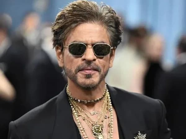 Shah Rukh Khan smiling on a red‑carpet, surrounded by flashing lights and a backdrop displaying his name in bold lettering.