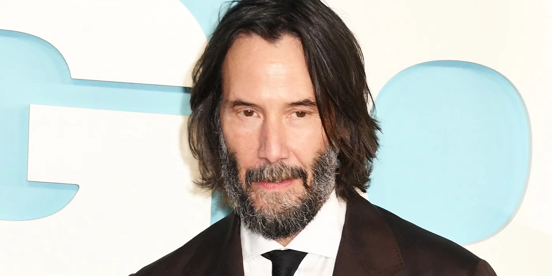 Keanu Reeves in a dark coat, standing in rain, looking ahead with focus.